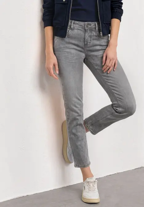 Eine Person steht in grauen, leicht ausgestellten Jeans und weißen Sneakern vor einer weißen Wand.