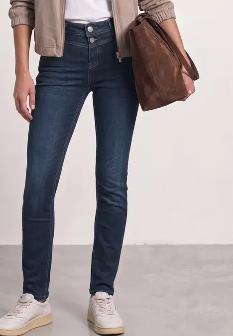 Eine Person trägt dunkelblaue Skinny Jeans, weiße Sneaker, eine beige Jacke und hält eine braune Tasche.