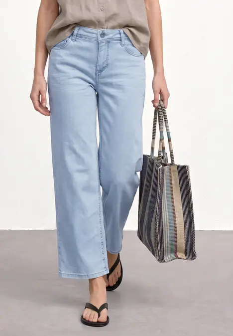 Eine Person trägt helle, weite Jeans und schwarze Flip-Flops, hält eine gestreifte Stofftasche.