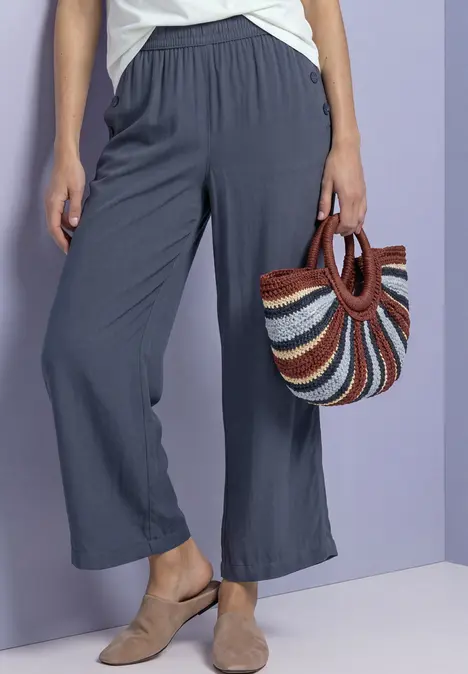 Frau trägt weite, graue Stoffhose und Pantoletten, hält eine bunte Strandtasche aus Stroh.