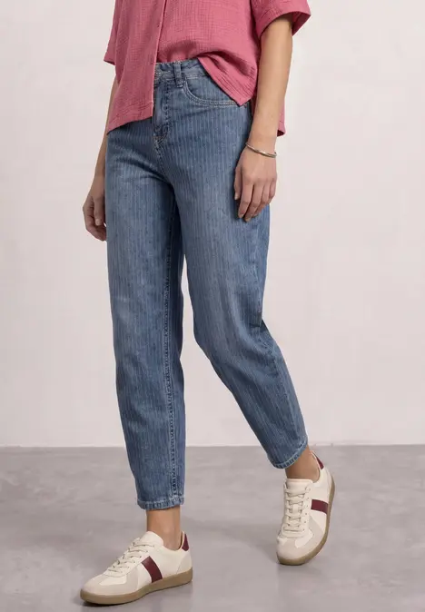 Frau in Jeans und pinkem Shirt, weiße Sneaker mit roten Streifen, stehend vor neutralem Hintergrund.