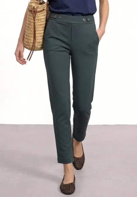 Eine Person trägt eine schmale, dunkelgrüne Stoffhose, hält eine geflochtene Basttasche und trägt gemusterte Ballerinas.