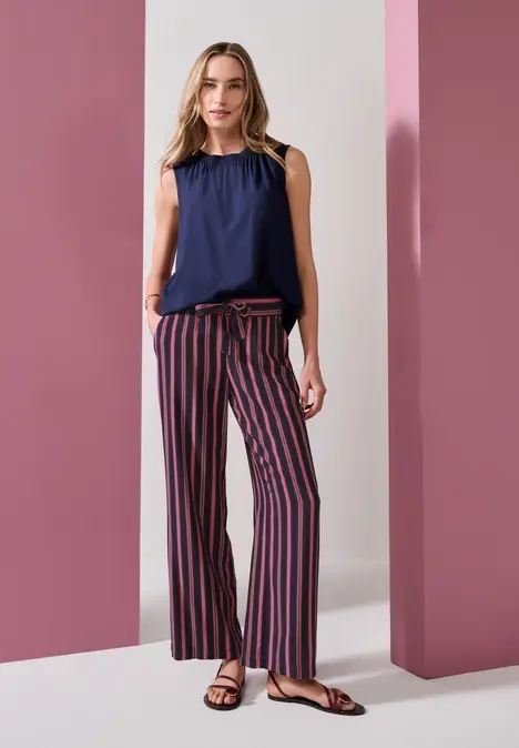 Frau in navy-blauer ärmelloser Bluse und gestreifter Marlene-Hose vor rosa Säulen.