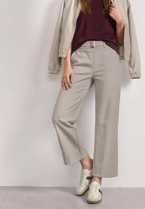 Person trägt beiges Culotte-Hose, bordeauxrotes Top, beigen Blazer und weiße Sneaker vor weißem Hintergrund.