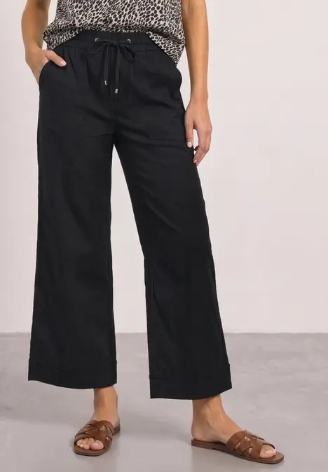 Frau in weiter schwarzer Leinenhose mit Kordelzug und braunen Sandalen, dazu gemustertes Top.