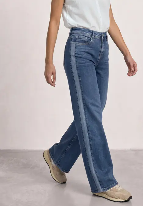 Eine Person trägt hochgeschnittene blaue Jeans mit seitlichem Streifen, ein weißes Top und robuste Stiefel.