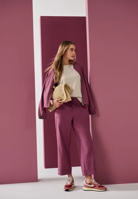 Frau in modernem pink-lila Hosenanzug mit beigem Oberteil und Sneakers posiert vor rosa Wand.