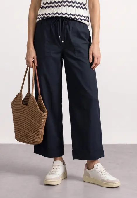 Person trägt dunkelblaue weite Hose, gestreiftes Top, weiße Sneaker und braune geflochtene Tasche.