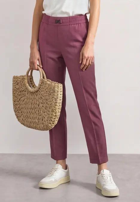 Person trägt rosafarbene Hose, weiße Sneaker und eine geflochtene Strohtasche in der Hand.
