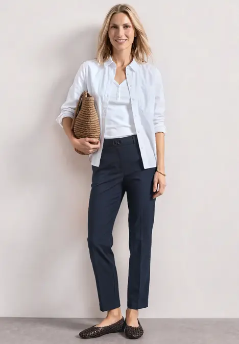 Eine Frau in Business-Casual-Outfit: weiße Bluse, weißer Cardigan, dunkelblaue Chinos, Loafers und Strohtasche.