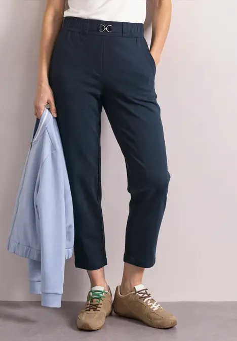 Person trägt taillierte, knöchellange dunkelblaue Hose, weißes Oberteil, helle Sneakers und hält eine hellblaue Jacke.