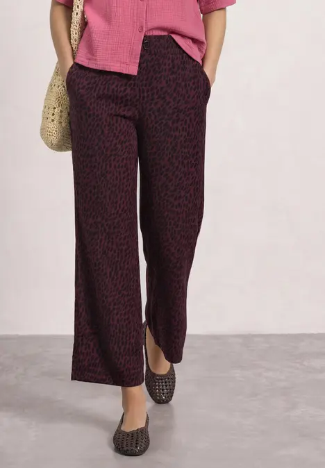 Frau trägt weite, dunkel gemusterte Hose, rosa Bluse, schwarze Ballerinas und Strohtasche.