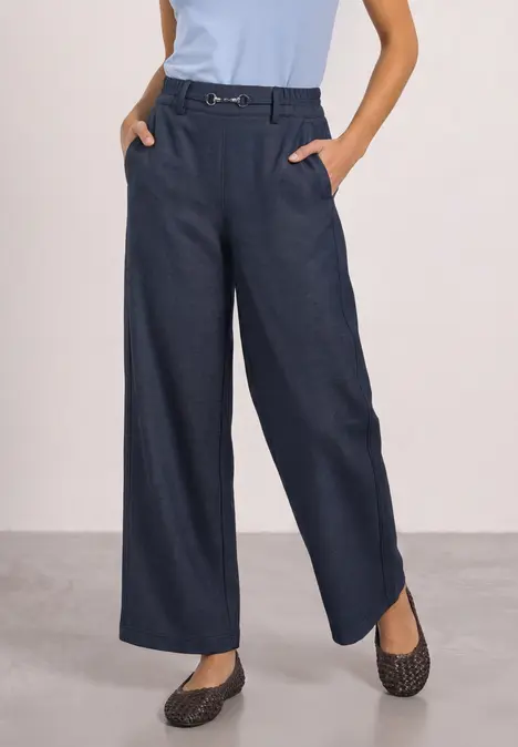Frau trägt dunkelblaue, weite Hose mit Gürtel, hellblaues Top und gemusterte Hausschuhe.