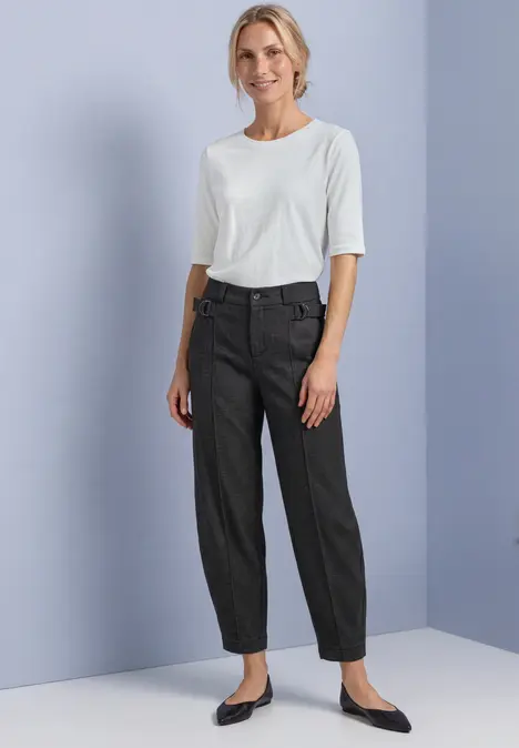Eine Frau steht in einer schlichten Umgebung, trägt ein weißes T-Shirt, graue Hose und schwarze Ballerinas.