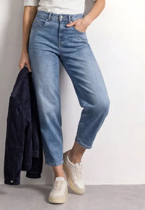 Persoon draagt lichtblauwe jeans, witte sneakers en houdt een donkere jas vast.
