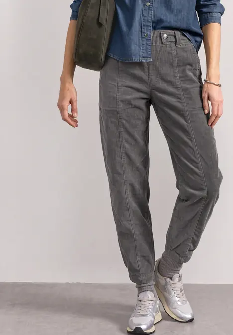 Unterkörper einer Person in blauer Jeansbluse, grauen Joggerhosen mit feinem Karomuster und weißen Sneakern.
