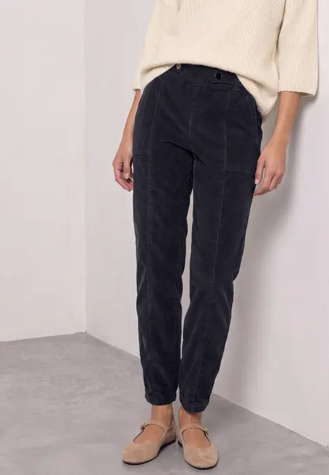 Schwarze, taillierte Cordhose mit schmalem Beinabschluss, kombiniert mit hellem Oberteil und Sandalen.