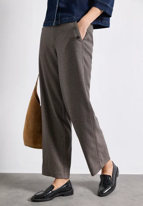 Person trägt graue Stoffhose, schwarze Loafers und hält eine braune Tasche.