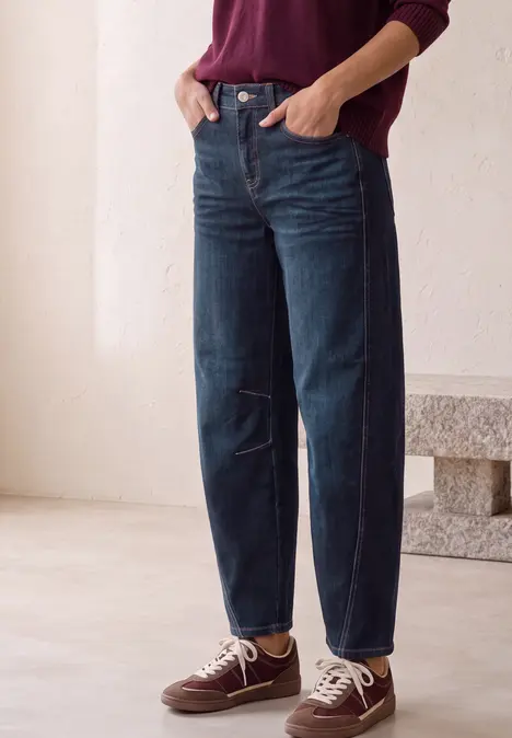 Person steht vor hellem Hintergrund in dunkelblauen Jeans, weinrotem Pullover und Sneakern.