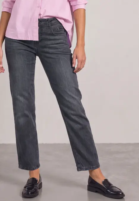 Unterer Teil einer Person in dunkelgrauen Straight-Jeans, schwarzen Loafern und rosafarbener Bluse vor neutralem Hintergrund.