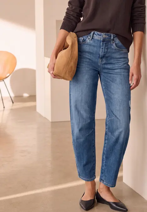 Frau trägt gerade geschnittene blaue Jeans, schwarze Ballerinas und beige Clutch vor minimalistischem Interieur.
