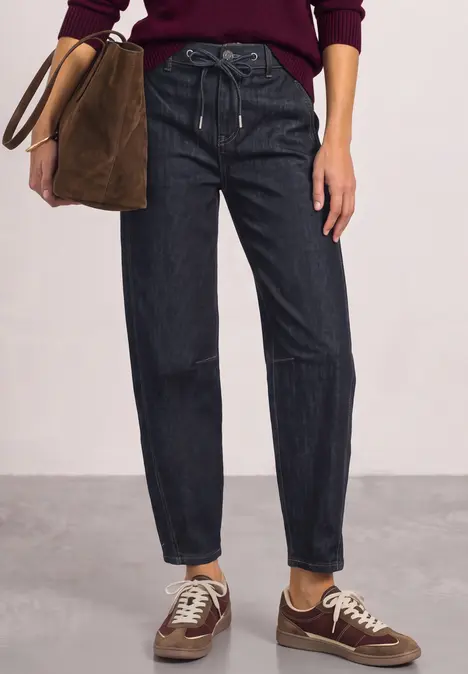 Frau in dunklen, lässigen Jeans mit Kordel, trägt braune Sneakers und hält eine Wildledertasche.