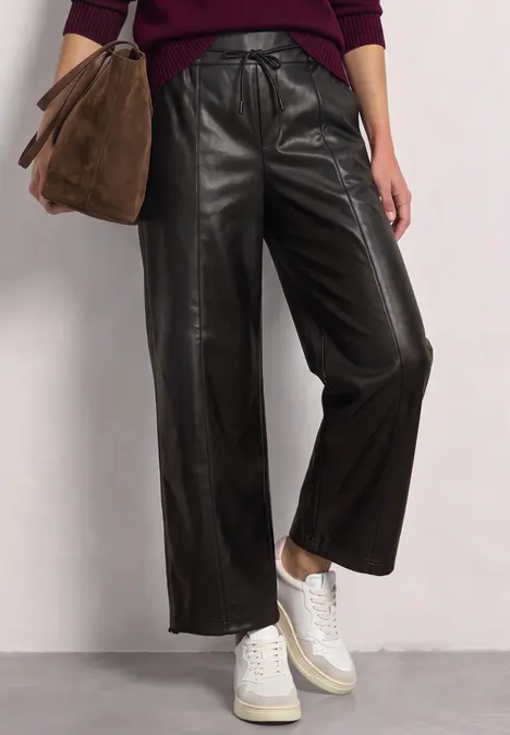 Modischer Look: schwarze, weite Lederhose, weiße Sneaker und braune Wildledertasche vor neutralem Hintergrund.