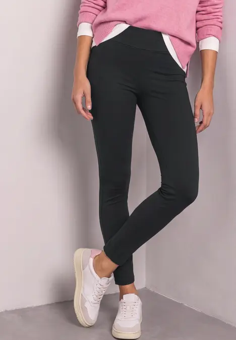 Frau in schwarzer Leggings, rosa Pullover und weißen Sneakern steht vor hellgrauer Wand.