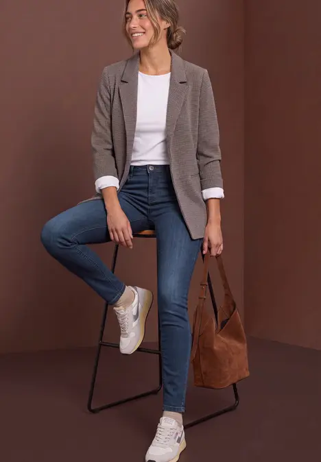 Frau in grauem Blazer, weißem T-Shirt, blauen Jeans und Sneakern, sitzt auf Hocker und hält braune Tasche.