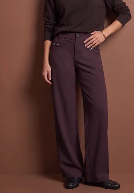 Femme en pantalon large violet foncé, haut sombre et chaussures noires sur fond marron.