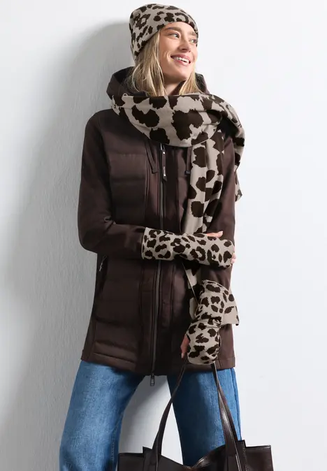 Frau lächelnd in brauner Winterjacke, Leo-Mütze, Schal und Handschuhen, hält Tasche.