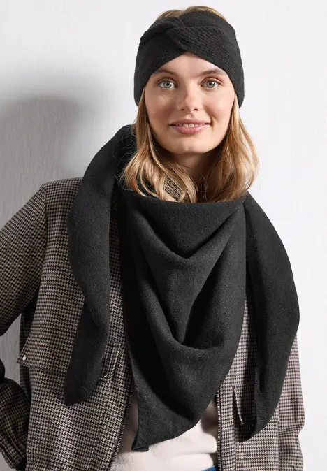 Frau mit schwarzem Stirnband und Dreieckstuch in schickem Karomantel, lächelnd vor heller Wand.