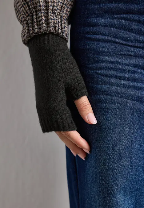 Schwarzer fingerloser Handschuh an einer Hand, kombiniert mit Jeans und Strickärmel im Hintergrund.