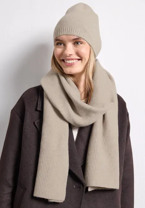 Femme souriante avec bonnet et écharpe en maille beige, manteau marron, fond blanc