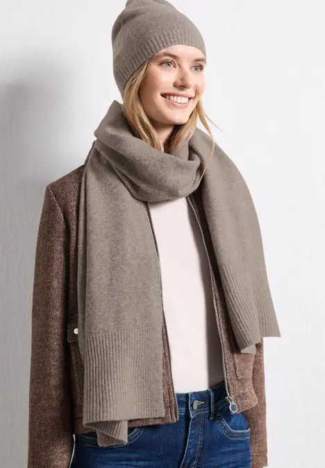 Frau in beiger Mütze und Schal, brauner Strickjacke und Jeans, lächelnd vor weißer Wand.