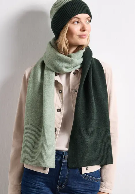 Frau mit grüner Strickmütze und zweifarbigem Schal trägt helle Jacke und Jeans, Blick zur Seite.