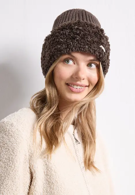 Junge Frau lächelt, trägt beige Teddyjacke und braune, flauschige Strickmütze.