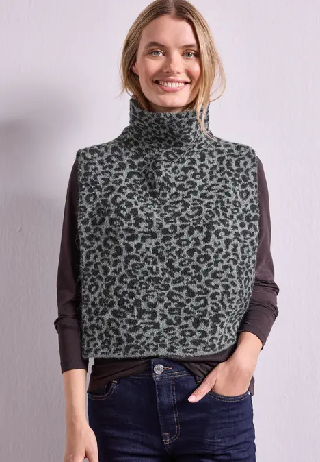 Vrouw draagt grijze leopard coltrui-vest over donkere longsleeve, glimlacht, handen in spijkerbroekzakken.