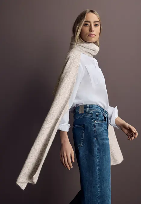 Vrouw in witte blouse, blauwe jeans en lange beige sjaal voor een grijze achtergrond.