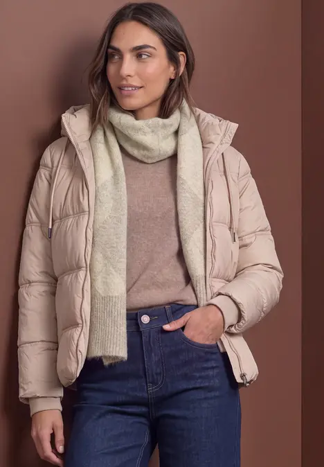 Eine Frau trägt eine beige Steppjacke, hellen Schal, Pullover und Jeans vor braunem Hintergrund.