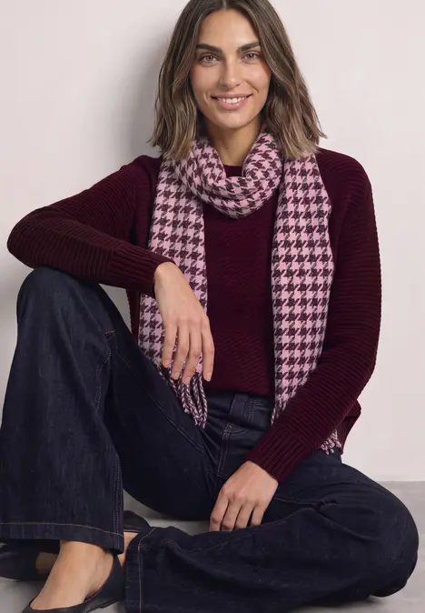 Frau in bordeauxrotem Pullover und karierter rosa Schal sitzt locker mit Jeans und lächelt.