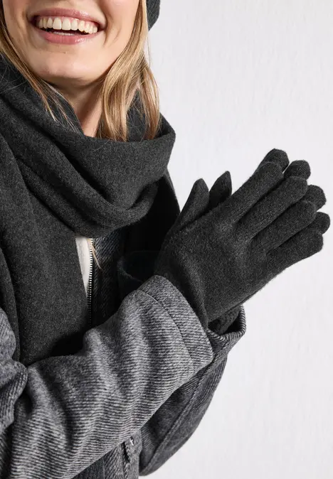 Lächelnde Person mit grauem Schal, grauem Mantel und schwarzen Handschuhen vor weißem Hintergrund.