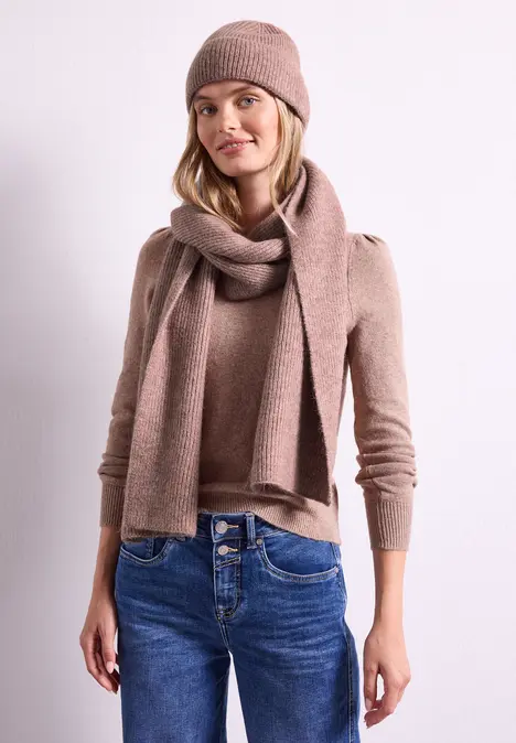 Jonge vrouw draagt beige gebreide muts, sjaal, trui en spijkerbroek voor een wit vlak.