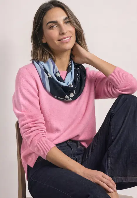 Frau in rosa Pullover und gemustertem Halstuch sitzt lässig auf einem Stuhl und lächelt leicht.