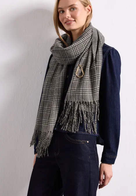 Modische Frau mit kariertem Fransen-Schal und dunklem Denim-Outfit vor weißer Wand.