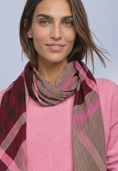 Eine Frau mit dunkelbraunen Haaren trägt einen rosa Pullover und einen gemusterten Schal, leicht lächelnd.