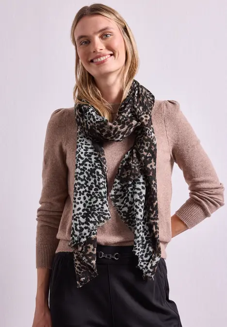 Eine Frau mit langem blondem Haar trägt braunen Pullover und gemusterten Schal, lächelnd vor weißem Hintergrund.