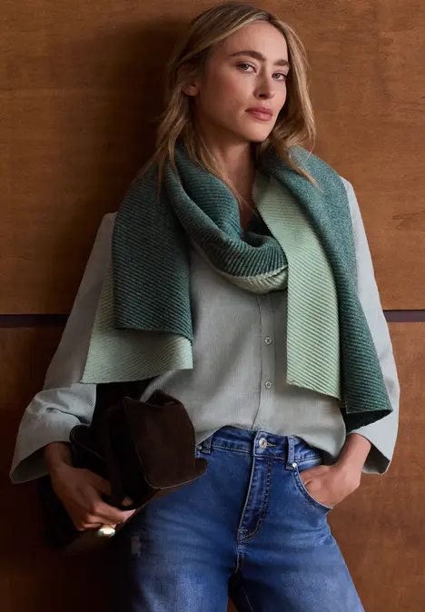 Vrouw in groene blouse en groene sjaal, blauwe jeans, houdt een bruine hoed vast voor een houten wand.