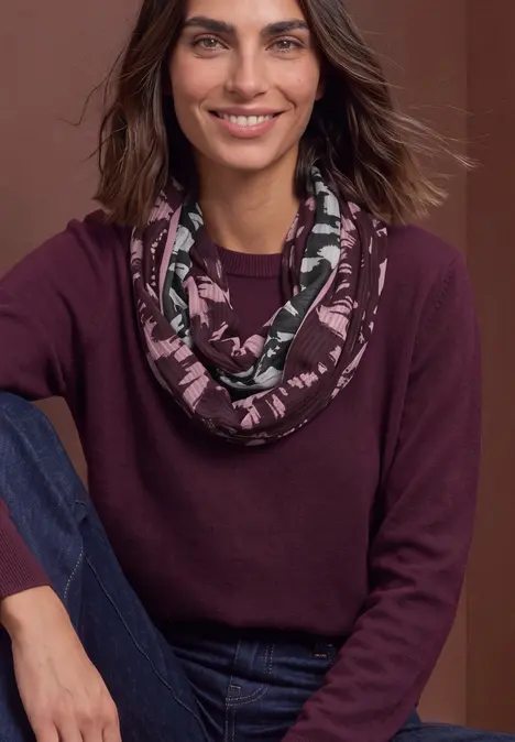 Frau mit dunkelrotem Pullover, gemustertem Loop-Schal und Jeans sitzt lächelnd.