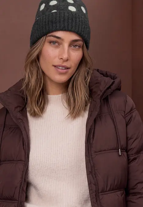 Femme en doudoune brune et bonnet à pois, fond marron.